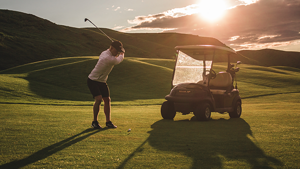 A golfer taking a swing with a golf cart in the background.