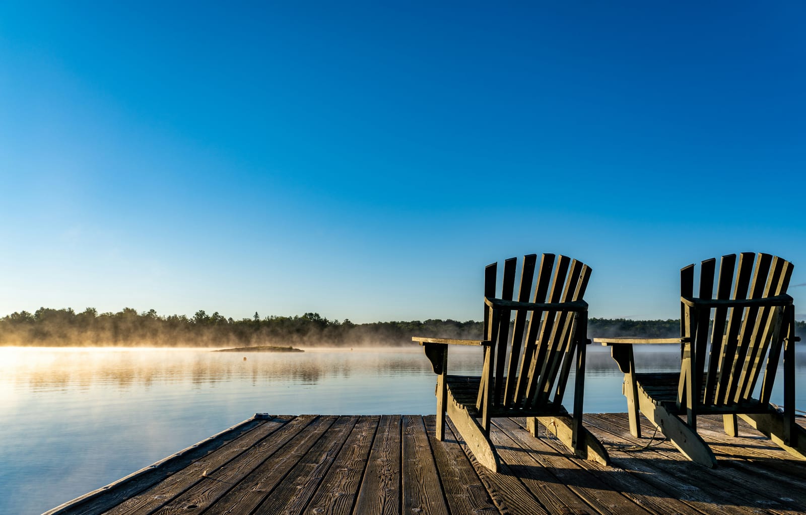 Two Adirondack chairs on a wooden dock overlooking a misty lake at sunrise
